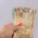 Mão segurando notas de real em destaque, representando pagamentos do programa Pé-de-Meia no novo ciclo de benefícios.