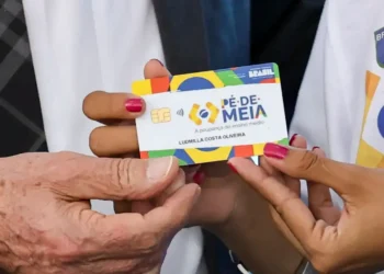 Mão segurando cartão do programa Pé-de-Meia durante liberação de pagamentos de R$ 1.000 no calendário de junho.