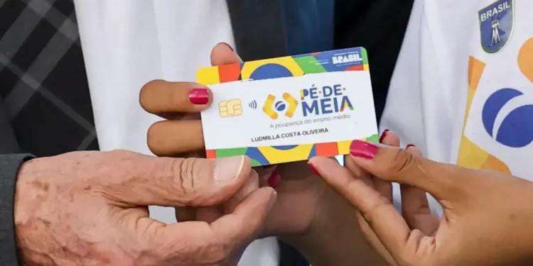 Mão segurando cartão do programa Pé-de-Meia durante liberação de pagamentos de R$ 1.000 no calendário de junho.
