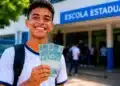 Estudante sorrindo em frente à escola segurando dinheiro, representando o pagamento do Pé-de-Meia de junho