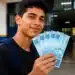 Jovem segurando cédulas de R$ 100, representando o pagamento do programa Pé-de-Meia, confirmado para junho, no valor de R$ 1.200.