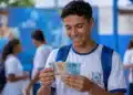 Estudante brasileiro de ensino médio segurando cédulas de reais em um pátio escolar, simbolizando recebimento de R$ 1.400 do Pé-de-Meia