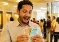 Trabalhador brasileiro segurando notas de reais com expressão de surpresa positiva em agência bancária, representando o pagamento do PIS/Pasep de R$ 4.151 em junho.