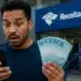 Homem surpreso consultando celular e segurando dinheiro em frente a prédio público, representando o Pix de R$ 1.000 da Receita Federal