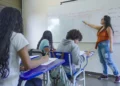 Professora explica conteúdo no quadro para estudantes em sala de aula da rede pública.