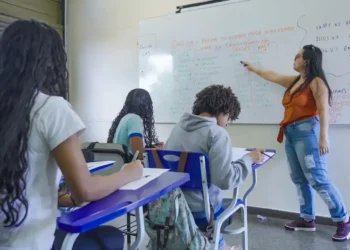 Professora explica conteúdo no quadro para estudantes em sala de aula da rede pública.