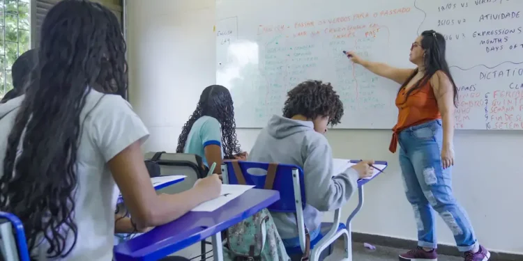 Professora explica conteúdo no quadro para estudantes em sala de aula da rede pública.