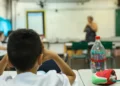 Aluno em sala de aula durante atividade escolar, com professora ao fundo, representando a Prova Nacional Docente 2026.