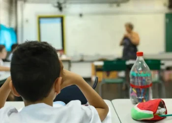 Aluno em sala de aula durante atividade escolar, com professora ao fundo, representando a Prova Nacional Docente 2026.