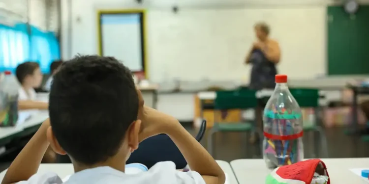 Aluno em sala de aula durante atividade escolar, com professora ao fundo, representando a Prova Nacional Docente 2026.