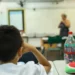 Aluno em sala de aula durante atividade escolar, com professora ao fundo, representando a Prova Nacional Docente 2026.