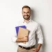 Homem sorridente segurando cadernos, representando estudante ou docente em contexto de preparação para a Prova Nacional Docente (PND).