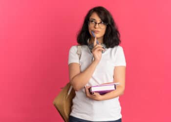 Estudante de óculos com caneta no queixo e livros na mão pensa em dúvida de português sobre fundo rosa
