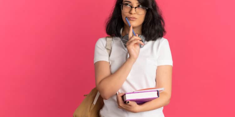 Estudante de óculos com caneta no queixo e livros na mão pensa em dúvida de português sobre fundo rosa