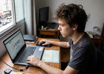 Jovem branco com camiseta escura usa notebook exibindo ENEM, sentado à mesa próximo à janela.