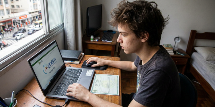 Jovem branco com camiseta escura usa notebook exibindo ENEM, sentado à mesa próximo à janela.