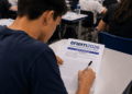 Jovem escreve à caneta no caderno de redação do ENEM 2026, em sala de aula lotada.