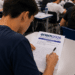 Jovem escreve à caneta no caderno de redação do ENEM 2026, em sala de aula lotada.