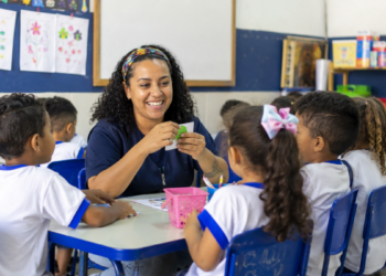 Professora orienta crianças em sala de aula de escola pública, representando vagas na área da Educação.