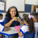 Professora orienta crianças em sala de aula de escola pública, representando vagas na área da Educação.