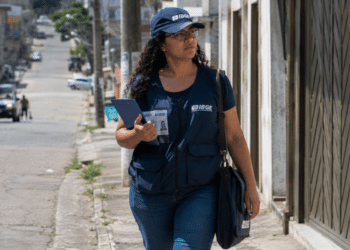 Agente do IBGE em trabalho de campo caminhando em rua urbana com prancheta.