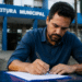 Homem de camisa azul escura assina documento com caneta preta, sentado em mesa externa diante de prédio da Prefeitura Municipal.