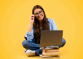 Jovem mulher de cabelo longo usando óculos e sorrindo, sentada com notebook sobre pilha de livros em fundo amarelo.