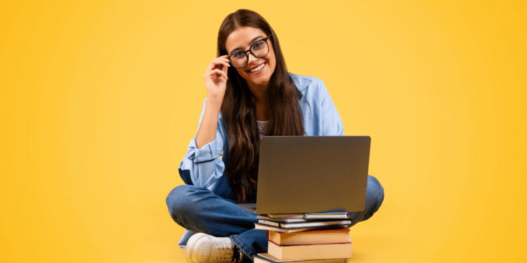 Jovem mulher de cabelo longo usando óculos e sorrindo, sentada com notebook sobre pilha de livros em fundo amarelo.