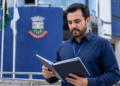 Homem de barba e crachá azul lendo pasta preta diante de prédio institucional com bandeiras.