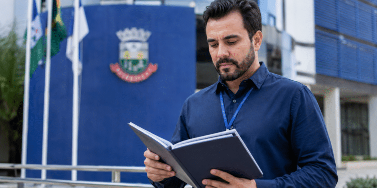 Homem de barba e crachá azul lendo pasta preta diante de prédio institucional com bandeiras.