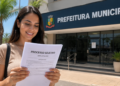 Mulher lendo edital de processo seletivo em frente à Prefeitura Municipal em dia ensolarado.