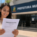 Mulher lendo edital de processo seletivo em frente à Prefeitura Municipal em dia ensolarado.