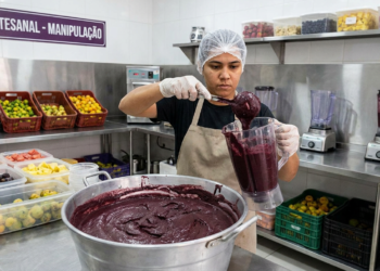 Pessoa manipulando mistura de açaí em cozinha industrial, cercada por frutas e utensílios.
