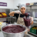 Pessoa manipulando mistura de açaí em cozinha industrial, cercada por frutas e utensílios.