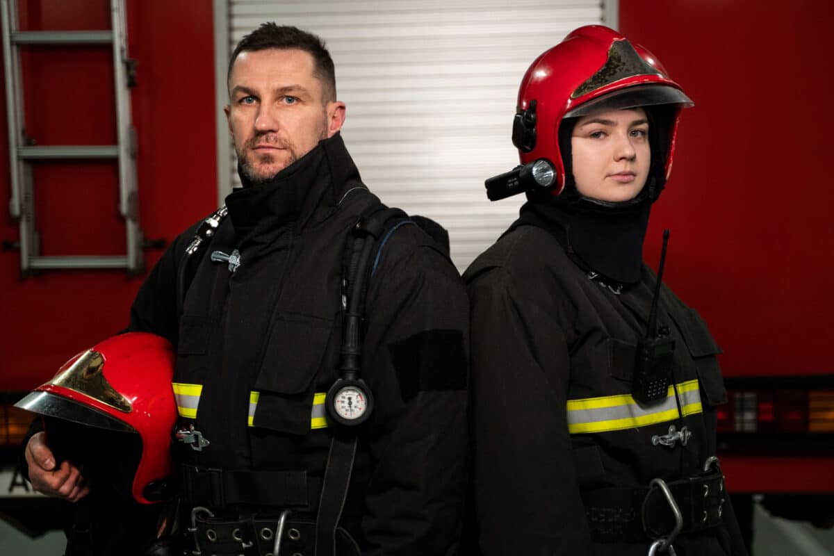 Dois bombeiros uniformizados com equipamento de combate a incêndio posam lado a lado diante de viatura vermelha