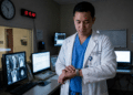 Homem asiático de jaleco branco observa relógio de pulso em sala com vários monitores de imagens médicas.