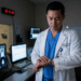 Homem asiático de jaleco branco observa relógio de pulso em sala com vários monitores de imagens médicas.