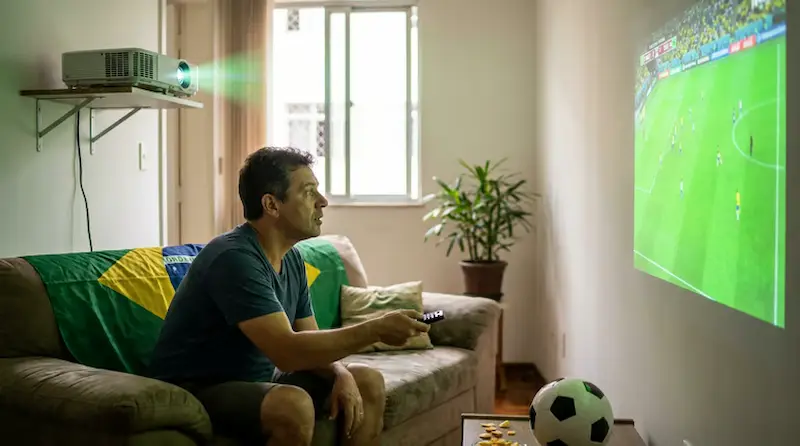 Pessoa sentada no sofá assiste a jogo de futebol projetado na parede com a tela posicionada à frente.