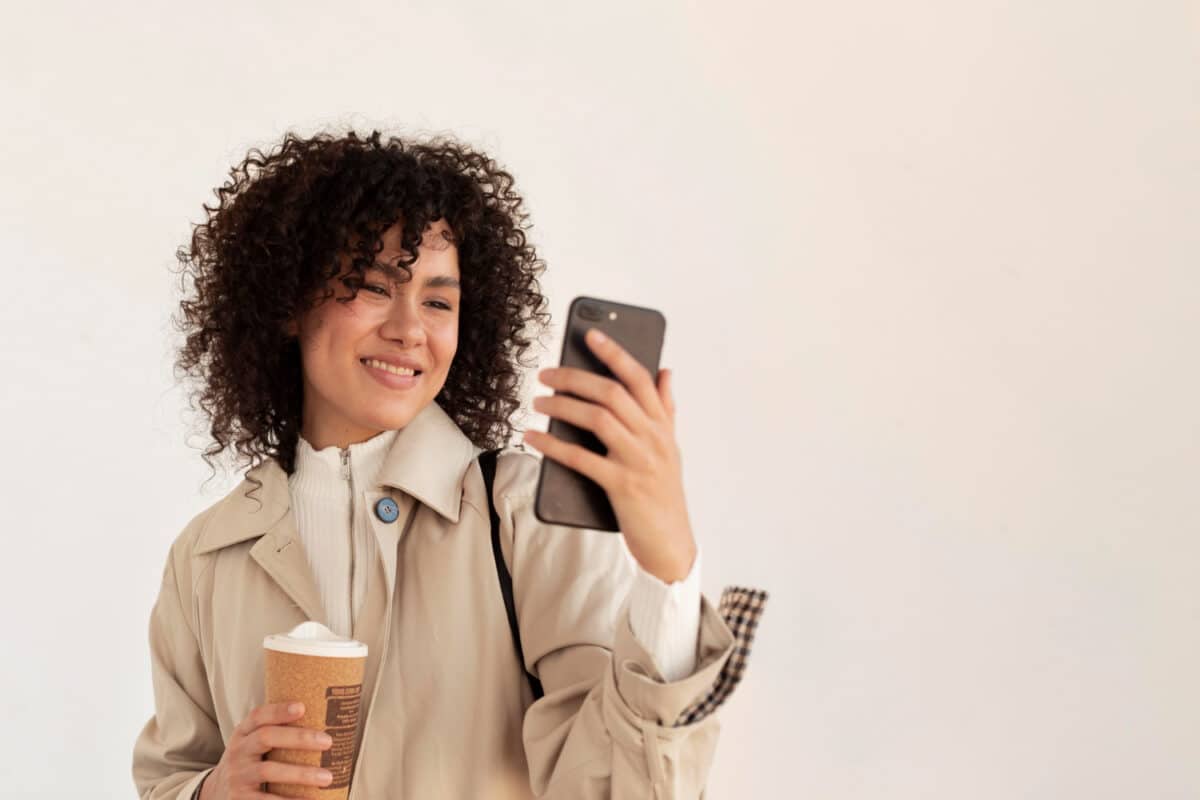 Mulher sorri com copo de café na mão enquanto observa o celular ao ar livre