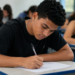 Estudante escrevendo redação do Enem em sala de aula com outros candidatos ao fundo.