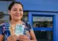 Mulher brasileira segurando cédulas de real em frente a uma agência bancária, simbolizando o pagamento da restituição do Imposto de Renda liberado pela Receita Federal.