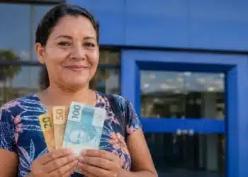 Mulher brasileira segurando cédulas de real em frente a uma agência bancária, simbolizando o pagamento da restituição do Imposto de Renda liberado pela Receita Federal.