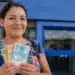 Mulher brasileira segurando cédulas de real em frente a uma agência bancária, simbolizando o pagamento da restituição do Imposto de Renda liberado pela Receita Federal.