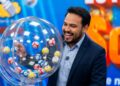 Homem de terno azul-marinho e camisa azul sorri ao lado de globo com bolas numeradas em sorteio de loteria