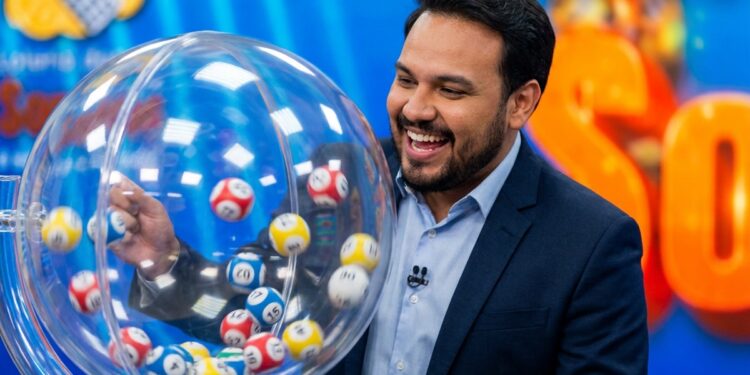 Homem de terno azul-marinho e camisa azul sorri ao lado de globo com bolas numeradas em sorteio de loteria