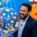 Homem de terno azul-marinho e camisa azul sorri ao lado de globo com bolas numeradas em sorteio de loteria