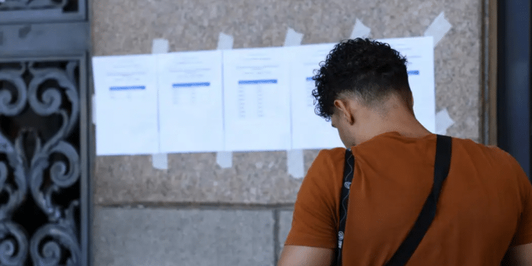 Jovem de camiseta laranja e alça de bolsa preta olha lista de aprovados fixada em parede de pedra.