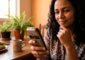 Mulher utiliza celular básico em mesa de cozinha com expressão de descoberta, em ambiente familiar e iluminado