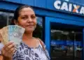 Mulher segura cédulas de real em frente a uma agência da Caixa, representando saque de R$ 2.900 disponível para CPFs com finais de 0 a 9.