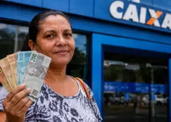 Mulher segura cédulas de real em frente a uma agência da Caixa, representando saque de R$ 2.900 disponível para CPFs com finais de 0 a 9.